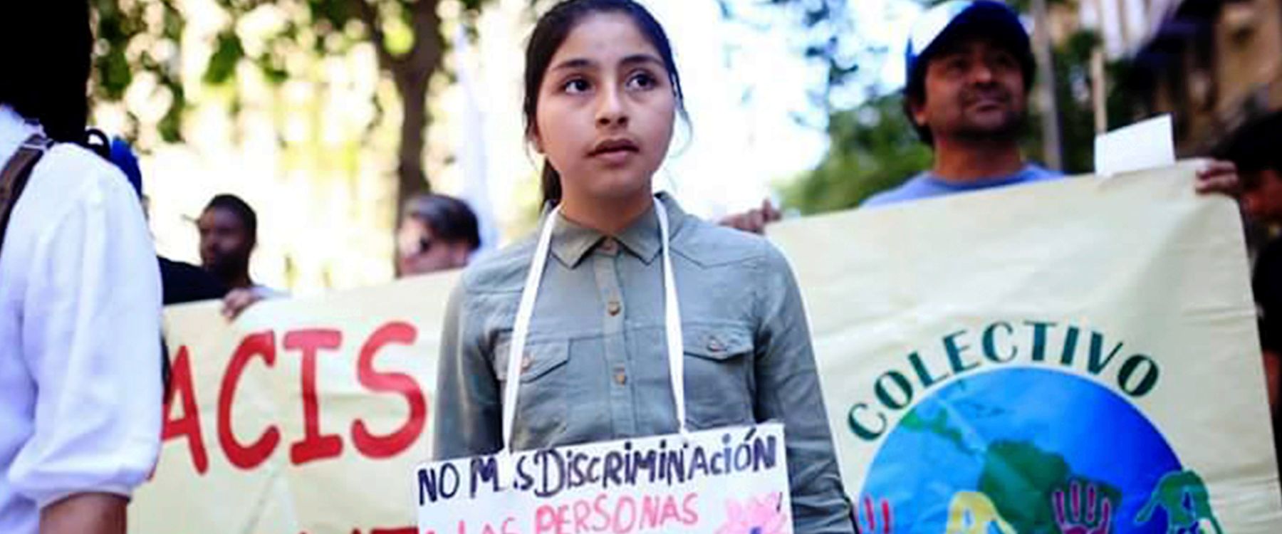 Una niña sostiene un cartel contra la discriminación en una marcha, junto al lienzo del Colectivo
