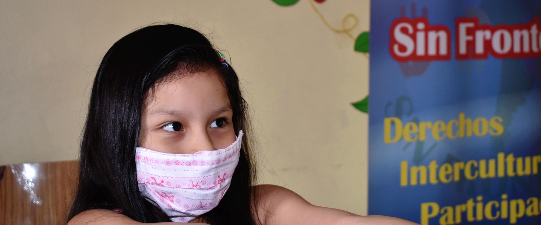 Una niña con mascarilla junto a un lienzo de Sin Fronteras que dice Derechos, Interculturalidad, Participación